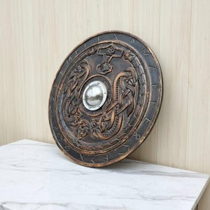 May include: A round, bronze-colored shield with intricate carvings of serpents, a hammer, and runes. A silver metal boss is in the center. The shield is displayed on a white marble surface.