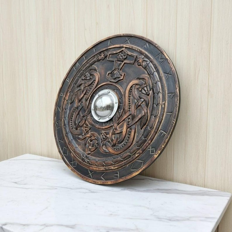 May include: A round, bronze-colored shield with intricate carvings of serpents, a hammer, and runes. A silver metal boss is in the center. The shield is displayed on a white marble surface.