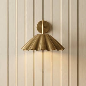 May include: A brass wall sconce featuring a circular mounting plate and a wide, downward-facing shade with a scalloped, fluted edge. The metal surface has a smooth, brushed finish that reflects light, creating a warm, metallic glow.