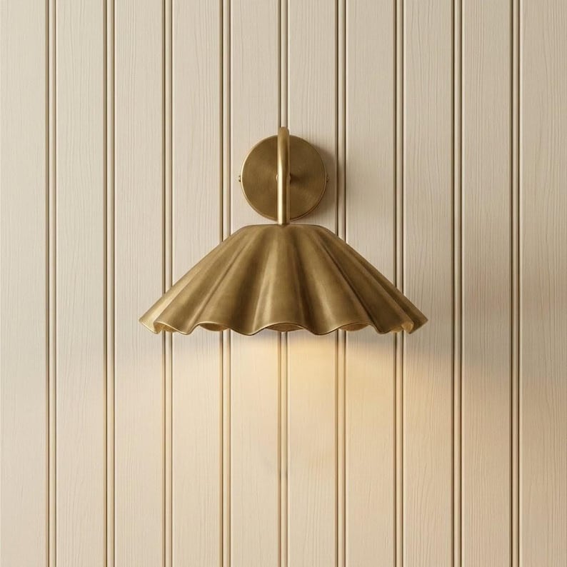 May include: A brass wall sconce featuring a circular mounting plate and a wide, downward-facing shade with a scalloped, fluted edge. The metal surface has a smooth, brushed finish that reflects light, creating a warm, metallic glow.