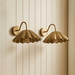 May include: A pair of wall-mounted lamps featuring brass-toned, scalloped, wide-brimmed shades. Each lamp has a curved arm extending from a circular wall plate, with a single exposed light bulb centered beneath the fluted metal shade.