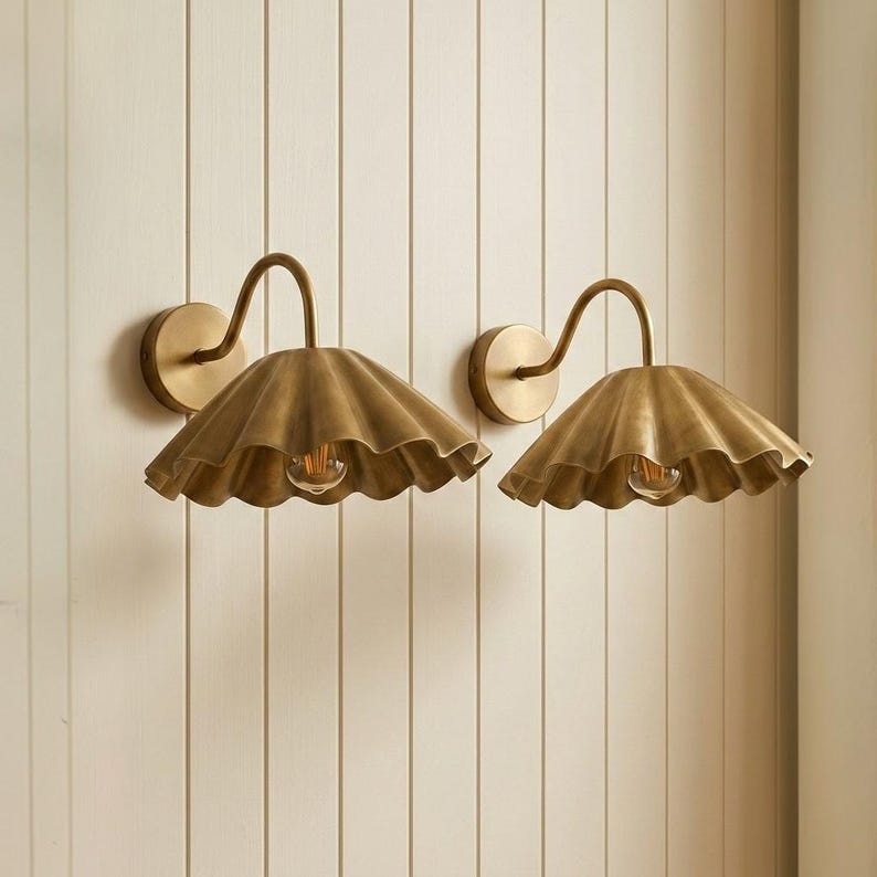 May include: A pair of wall-mounted lamps featuring brass-toned, scalloped, wide-brimmed shades. Each lamp has a curved arm extending from a circular wall plate, with a single exposed light bulb centered beneath the fluted metal shade.