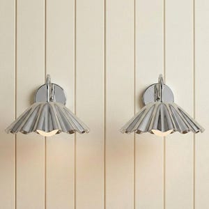 May include: A pair of wall-mounted light fixtures featuring polished chrome finishes. Each lamp has a circular backplate and a wide, fluted, fan-shaped shade that directs light downward, revealing a small, glowing bulb at the center of each.
