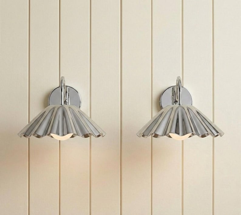 May include: A pair of wall-mounted light fixtures featuring polished chrome finishes. Each lamp has a circular backplate and a wide, fluted, fan-shaped shade that directs light downward, revealing a small, glowing bulb at the center of each.