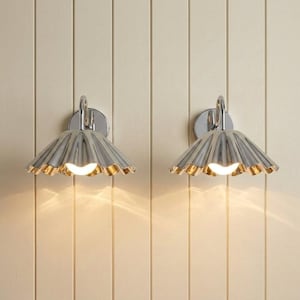 May include: A pair of wall-mounted light fixtures featuring polished chrome finishes. Each lamp has a wide, scalloped, fan-shaped shade with a reflective metallic surface and a single central light bulb that emits a warm, soft glow downward.