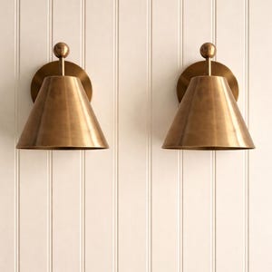 May include: Two brass wall sconces with conical shades and a round backplate are mounted on a white paneled wall. Each sconce has a small, spherical detail at the top. The sconces are made of brass and have a warm, metallic finish.