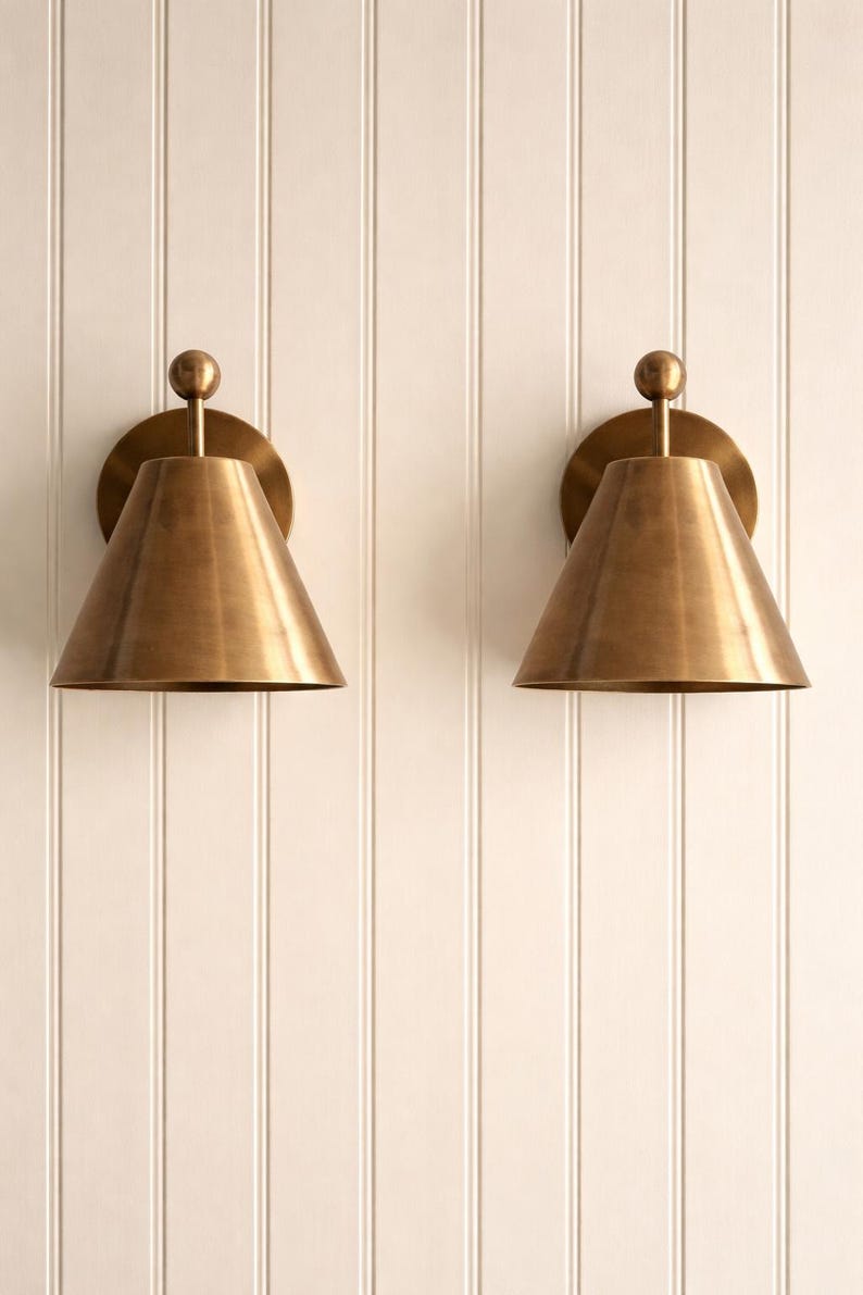 May include: Two brass wall sconces with conical shades and a round backplate are mounted on a white paneled wall. Each sconce has a small, spherical detail at the top. The sconces are made of brass and have a warm, metallic finish.