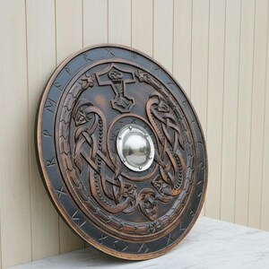 May include: A round, wooden shield with intricate carvings and a dark brown finish. The shield features a central metal boss and a border with runic inscriptions. The design includes Viking-style knotwork and a hammer symbol.