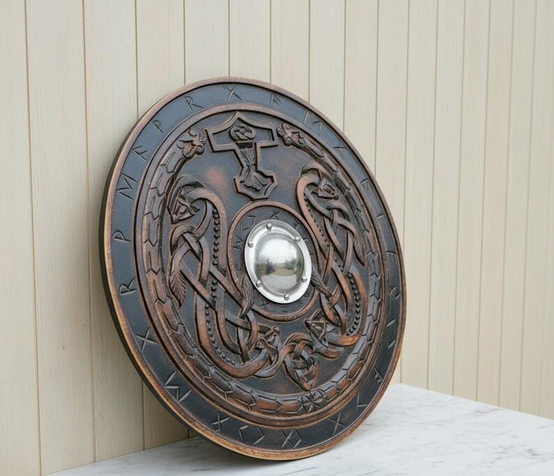 May include: A round, wooden shield with intricate carvings and a dark brown finish. The shield features a central metal boss and a border with runic inscriptions. The design includes Viking-style knotwork and a hammer symbol.