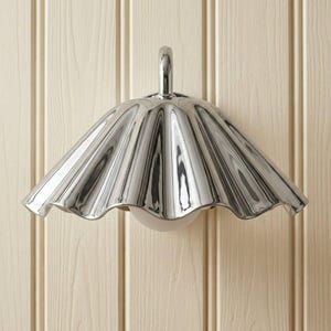 May include: A wall-mounted light fixture featuring a polished chrome, scalloped, fan-shaped shade. The metallic surface has a reflective finish with deep, vertical ridges that create a pleated appearance. A small, rounded white bulb is visible underneath.
