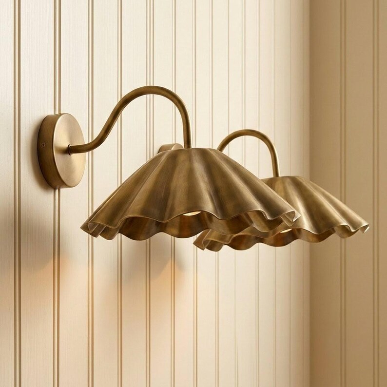 May include: A double wall sconce light fixture featuring two curved arms extending from a circular base. Each arm supports a wide, scalloped, brass-toned metal shade with a matte finish, directing light downward from the center of each shade.