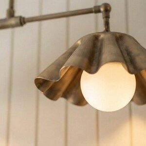May include: A brass-toned metal pendant light fixture featuring a scalloped, fluted shade design. The fixture includes a central, spherical, frosted white glass bulb that emits a soft, warm glow, suspended from a matching horizontal metal arm.