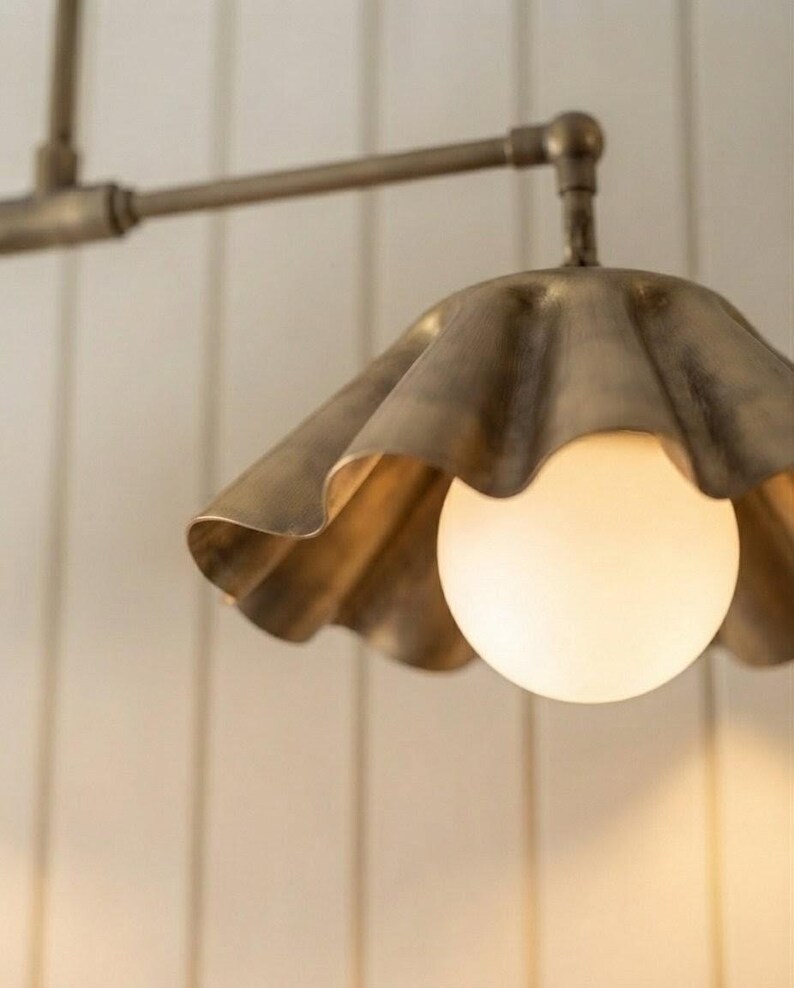 May include: A brass-toned metal pendant light fixture featuring a scalloped, fluted shade design. The fixture includes a central, spherical, frosted white glass bulb that emits a soft, warm glow, suspended from a matching horizontal metal arm.