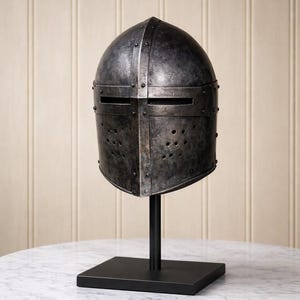 May include: A weathered, antique-style metal helmet with a riveted construction, displayed on a black stand. The helmet features eye slits and ventilation holes, showcasing a medieval aesthetic. The helmet is displayed on a black stand with a square base.
