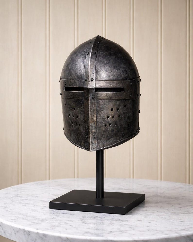 May include: A weathered, antique-style metal helmet with a riveted construction, displayed on a black stand. The helmet features eye slits and ventilation holes, showcasing a medieval aesthetic. The helmet is displayed on a black stand with a square base.