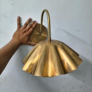 May include: A brass wall sconce featuring a curved arm and a wide, fluted, scalloped shade. The metal has a brushed, matte finish with a warm golden tone. The fixture includes a circular wall mounting plate and a downward-facing bell-shaped shade.