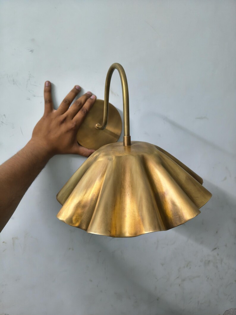 May include: A brass wall sconce featuring a curved arm and a wide, fluted, scalloped shade. The metal has a brushed, matte finish with a warm golden tone. The fixture includes a circular wall mounting plate and a downward-facing bell-shaped shade.