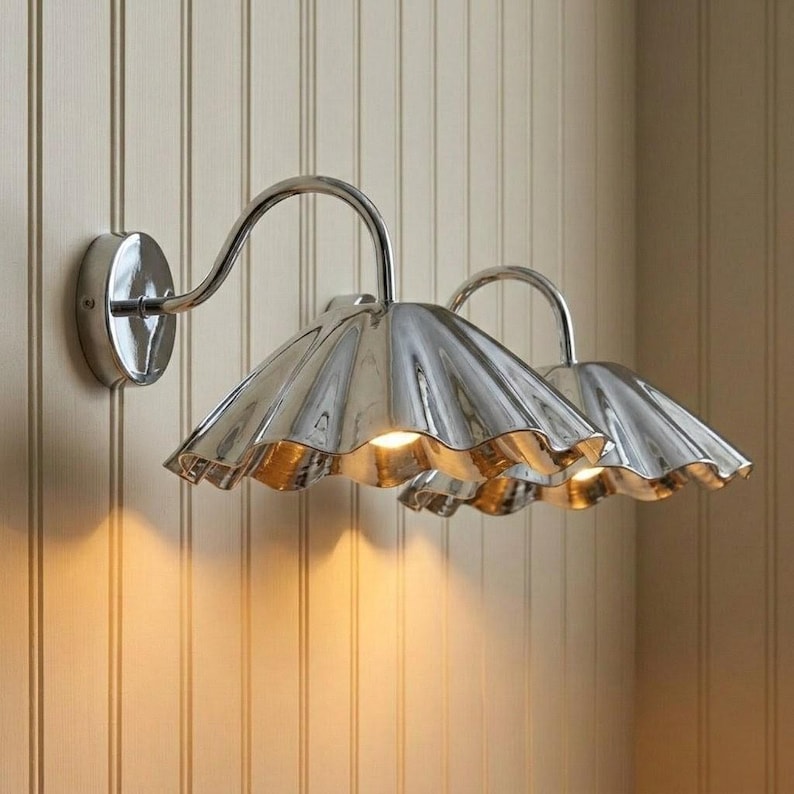 May include: A double wall sconce featuring two polished chrome fixtures with curved arms and wide, scalloped, bell-shaped shades. The metallic surfaces reflect light, and the design includes a circular mounting plate attached to the wall.
