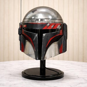 May include: A silver and black helmet with red detailing, displayed on a black stand. The helmet has a unique design with a T-shaped visor and a small aerial. The item is likely a prop or collectible.