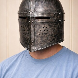 May include: A weathered, metallic medieval helmet with riveted construction and a dark, aged finish. The helmet features horizontal eye slits and ventilation holes. The person is wearing a light blue crew neck t-shirt.
