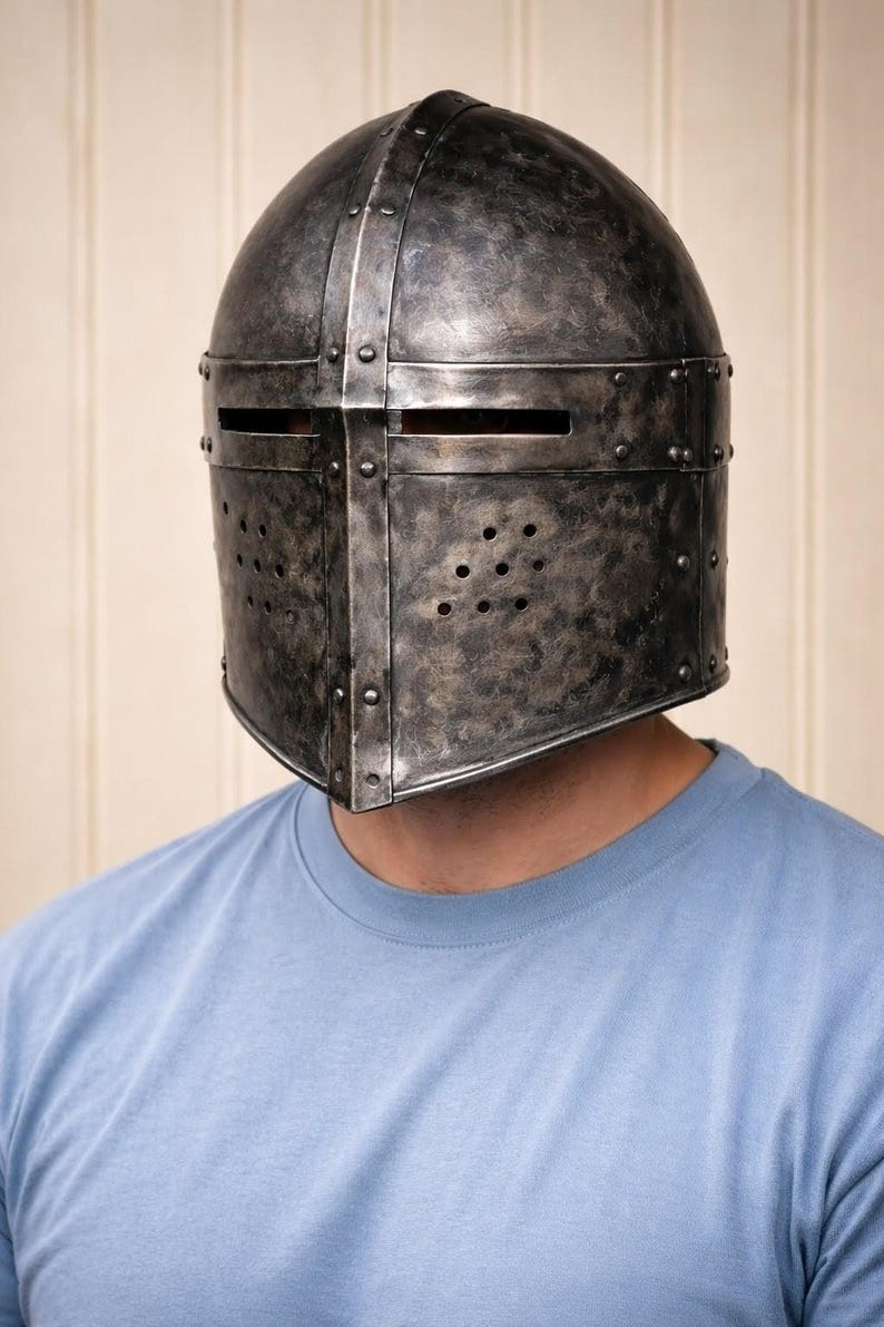 May include: A weathered, metallic medieval helmet with riveted construction and a dark, aged finish. The helmet features horizontal eye slits and ventilation holes. The person is wearing a light blue crew neck t-shirt.