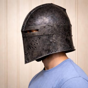 May include: A dark gray metal helmet with a riveted construction. The helmet features a visor with a horizontal eye slit and several small ventilation holes. The helmet is worn by a person wearing a blue t-shirt.
