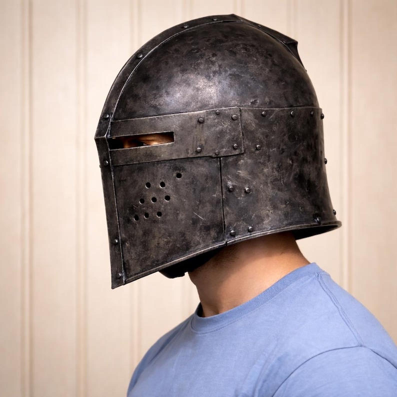May include: A dark gray metal helmet with a riveted construction. The helmet features a visor with a horizontal eye slit and several small ventilation holes. The helmet is worn by a person wearing a blue t-shirt.
