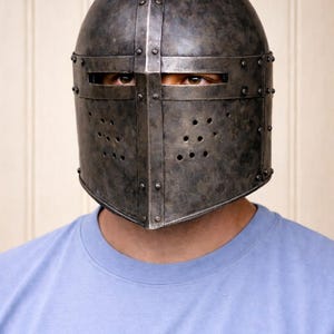 May include: A medieval-style metal helmet with a riveted construction and a dark, aged finish. The helmet features a visor with eye slits and ventilation holes. The person is wearing a light blue t-shirt.