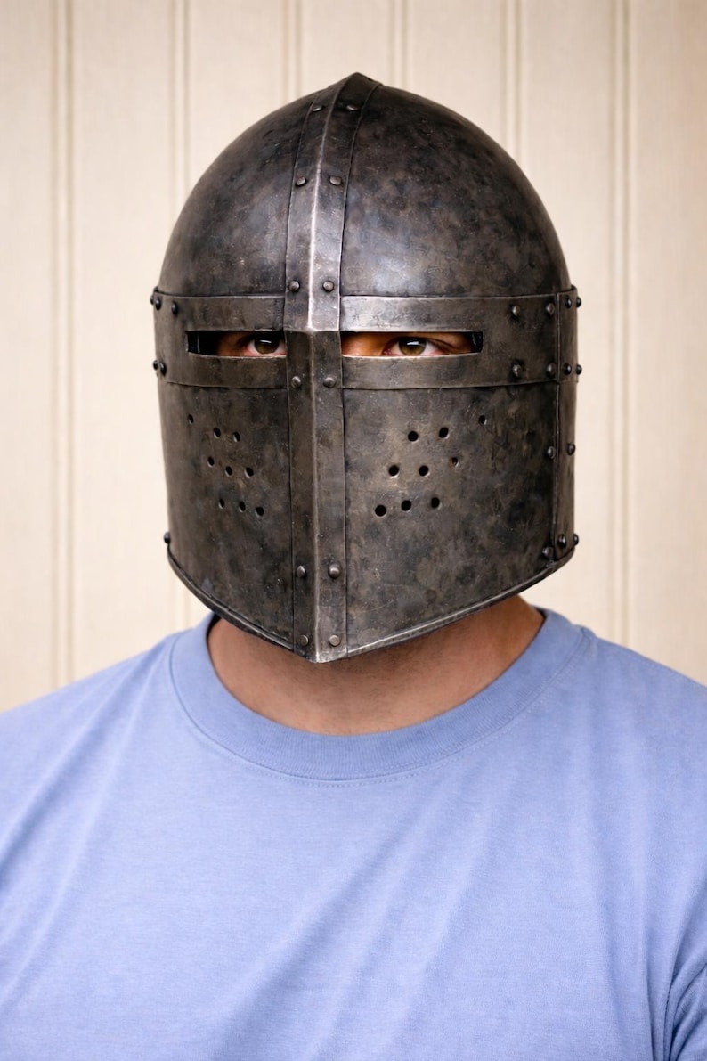 May include: A medieval-style metal helmet with a riveted construction and a dark, aged finish. The helmet features a visor with eye slits and ventilation holes. The person is wearing a light blue t-shirt.