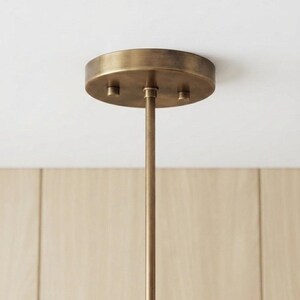 May include: A circular antique brass ceiling canopy with a matching thin vertical rod extending downward. The canopy features a brushed metallic finish and is secured with three small, evenly spaced cylindrical fasteners on its flat surface.