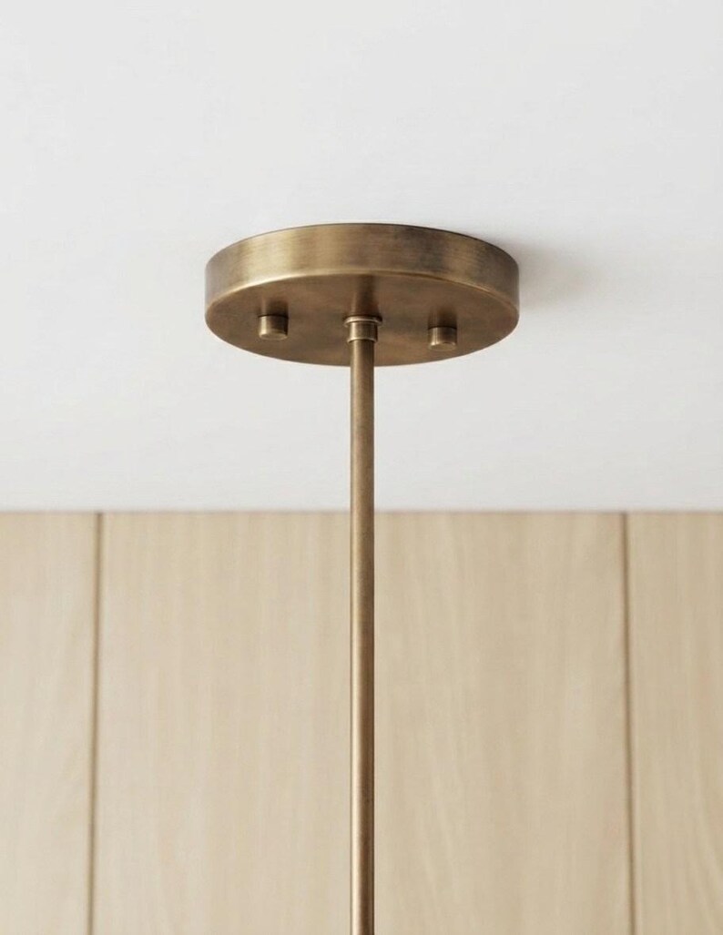 May include: A circular antique brass ceiling canopy with a matching thin vertical rod extending downward. The canopy features a brushed metallic finish and is secured with three small, evenly spaced cylindrical fasteners on its flat surface.