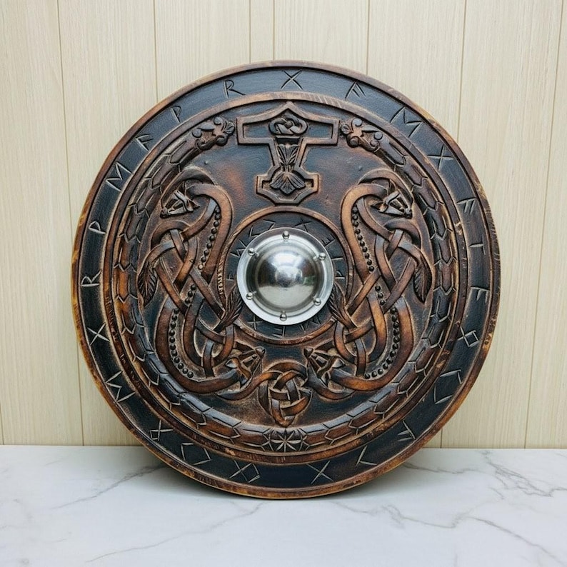 May include: A round, wooden shield with intricate carvings. The shield features a central metal boss and is decorated with Norse knotwork, serpents, and a hammer symbol. The shield is primarily brown with dark accents.