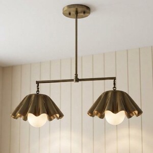 May include: A dual-light pendant fixture featuring a brass-toned metal frame with a central rod and horizontal bar. Two fluted, bell-shaped metal shades in a matching brass finish hang from the ends, each housing a round, frosted white glass bulb.