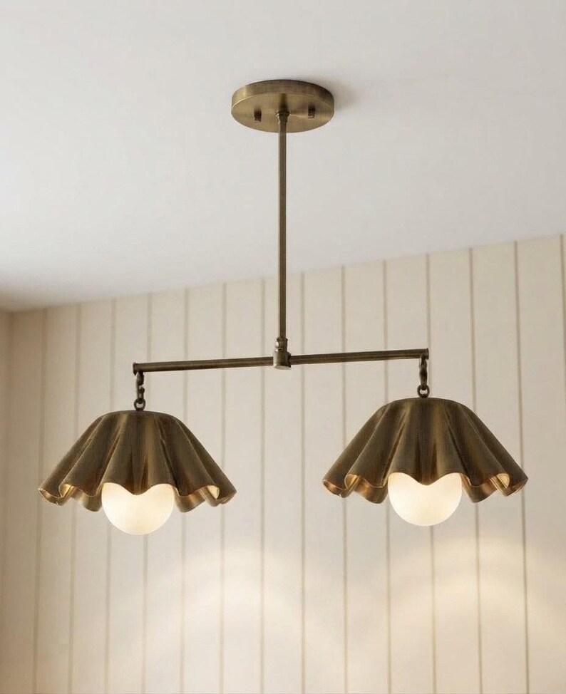 May include: A dual-light pendant fixture featuring a brass-toned metal frame with a central rod and horizontal bar. Two fluted, bell-shaped metal shades in a matching brass finish hang from the ends, each housing a round, frosted white glass bulb.