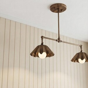 May include: A ceiling-mounted light fixture featuring a horizontal metal rod in a bronze finish, connected to a circular canopy. Two downward-facing, fluted metal shades in a matching bronze tone each house a single round white light bulb.