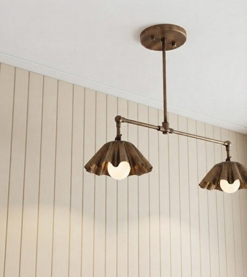 May include: A ceiling-mounted light fixture featuring a horizontal metal rod in a bronze finish, connected to a circular canopy. Two downward-facing, fluted metal shades in a matching bronze tone each house a single round white light bulb.