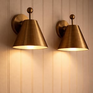 May include: Two brass-colored wall sconces with a conical shade and a small ball finial. The sconces are mounted on a light-colored, vertically-paneled wall. The sconces are lit, casting a warm glow.