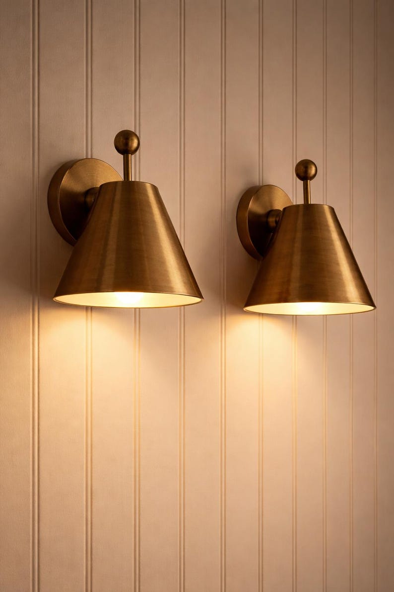 May include: Two brass-colored wall sconces with a conical shade and a small ball finial. The sconces are mounted on a light-colored, vertically-paneled wall. The sconces are lit, casting a warm glow.