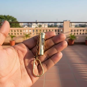May include: A brass hourglass keychain is held in a hand. The hourglass has a clear glass center. The keychain has a chain and a clasp. The background shows a rooftop with plants and a cityscape. The hourglass is approximately 6 cm in length.