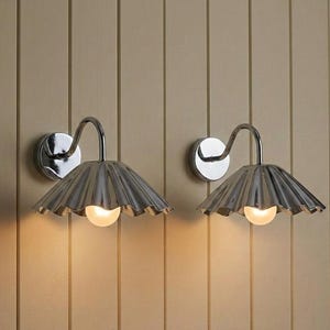 May include: A pair of wall-mounted lamps featuring polished chrome finishes, curved arms, and fluted, fan-shaped shades. Each lamp emits a warm, soft glow from a single light bulb positioned beneath the metallic, pleated shade structure.