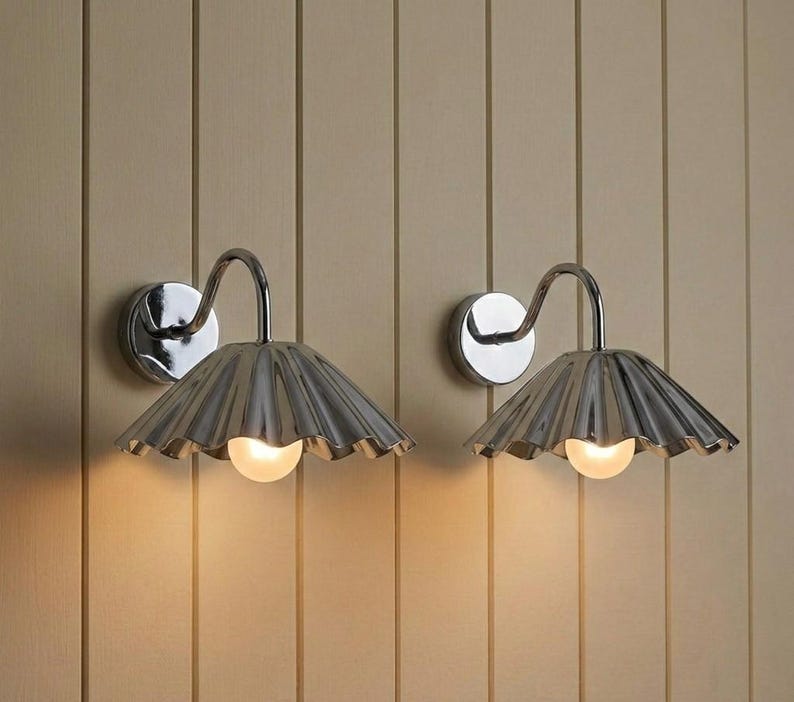 May include: A pair of wall-mounted lamps featuring polished chrome finishes, curved arms, and fluted, fan-shaped shades. Each lamp emits a warm, soft glow from a single light bulb positioned beneath the metallic, pleated shade structure.