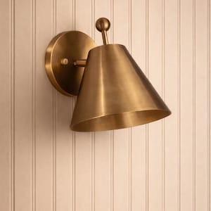 May include: A brass wall sconce with a conical shade and a round backplate. The sconce is mounted on a white vertical-paneled wall. The fixture has a small ball at the top.