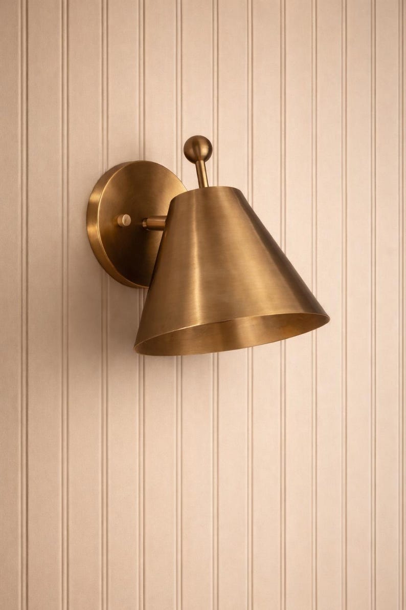 May include: A brass wall sconce with a conical shade and a round backplate. The sconce is mounted on a white vertical-paneled wall. The fixture has a small ball at the top.