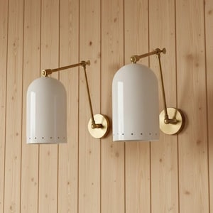 May include: Two wall-mounted lamps featuring white dome-shaped shades with a row of small perforations along the bottom edge. Each lamp has an adjustable brass-toned metal arm and a circular wall plate, providing a functional lighting solution.