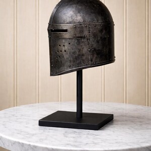 May include: A weathered, dark metal helmet with a visor slit and rivet details, displayed on a black stand. The helmet is set against a neutral background, showcasing its historical design. The stand has a square base and a vertical rod.
