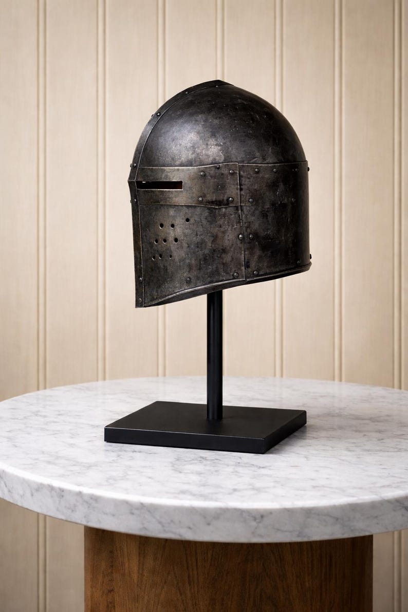 May include: A weathered, dark metal helmet with a visor slit and rivet details, displayed on a black stand. The helmet is set against a neutral background, showcasing its historical design. The stand has a square base and a vertical rod.