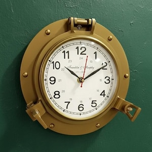 Può includere: Un orologio da parete color oro con design a oblò. Il quadrante è bianco con numeri e lancette nere, e il testo "Franklin & Murphy EST 184" è visibile. L'orologio è montato su una parete verde.