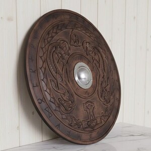 May include: A round, wooden shield with intricate carvings. The shield is a dark brown color and features a central metal boss. The design includes intertwined serpent-like figures and a hammer symbol, with runic inscriptions around the edge.