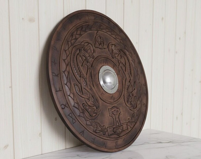 May include: A round, wooden shield with intricate carvings. The shield is a dark brown color and features a central metal boss. The design includes intertwined serpent-like figures and a hammer symbol, with runic inscriptions around the edge.