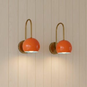 May include: Two orange wall sconces with a brass finish. Each sconce has a rounded shade and a curved arm. The sconces are mounted on a light-coloured wooden wall. The interior of the shade is white.